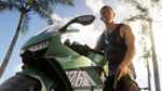 Man in a black tank top and sunglasses beside a green sport motorcycle under palm trees the sunny sky behind.