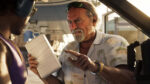 Older man in tropical shirt hands a wrapped brick to a younger man in a car, pointing and speaking intensively.