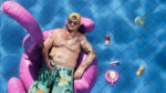 Shirtless tattooed man wearing sunglasses and a yellow cap lounges on a pink inflatable flamingo in a blue tiled pool, holding a beer can.