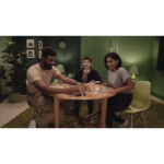 Three friends play a board game around a round wooden table in a cozy green-lit room, smiling and focused on the game.