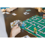 515V7_tO8-L_AC_SL1024___324301785270583 - Video Game Depot People playing a board game with cards in hand and a green maze-like board on a wooden table.