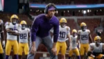 517f965c-3de7-43c7-b900-280f60b5ebe2 - Video Game Depot LSU coach in purple shirt and headset crouches on the field, with players in yellow pants behind him during a game.