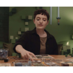 Person with short dark hair sits at a table playing a board game, reaching to place a card on the board.
