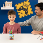 Boy sits at a white table with colored pencils in a Mario-themed cup, smiling while an adult sits beside him in a room with a yellow 'GAMER ZONE' sign on a blue wall.