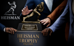 6383fe5b-25d1-4821-8ea6-307ba5718f5d - Video Game Depot Hands rest on a large Heisman Trophy statue on a pedestal, with 'HEISMAN TROPHY EST.1935' text visible behind.