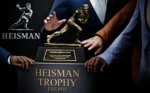 6383fe5b-25d1-4821-8ea6-307ba5718f5d - Video Game Depot Bronze Heisman Trophy statue on a dark pedestal, hands of presenters around it during the award ceremony.