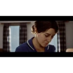 Woman in a blue uniform looks down, indoors with plaid curtains in the background, soft lighting.