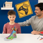 71wImuZMO5L_AC_SL1500___568431785270348 - Video Game Depot Boy and man sit at a table with a colorful high-top sneaker filled with scissors and markers on the table, blue wall in background with a Gamer Zone sign.