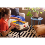 81V4Jc_wBaL_AC_SL1500___672751785344560 - Video Game Depot Young girl in a red sweater plays a wooden Pac-Man tabletop game on a small round table in a cozy living room with plants and cushions nearby.