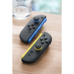 Two Nintendo Switch Joy-Con controllers (blue and yellow) attached to a charging grip on a wooden surface beside a glass.