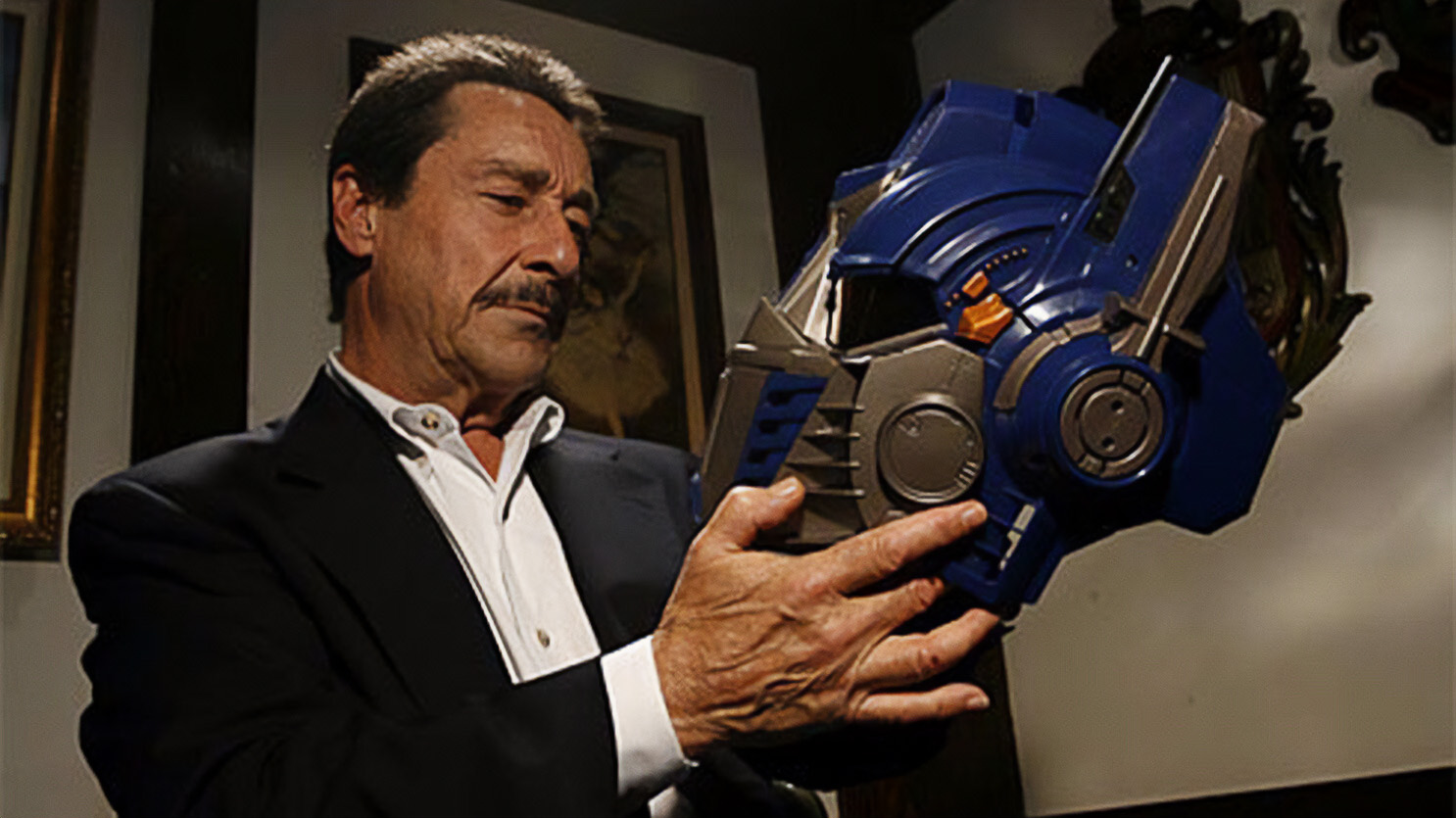 Man in a suit holding a large blue robot head helmet, inspecting it intently by a framed painting in the background